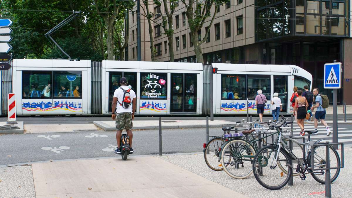 Une rame de tramway, des piétons sur un passage piéton, des vélos stationnés et une personne sur un onewheel sur une piste cyclable.